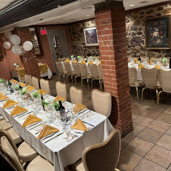 Long dining tables set with white tablecloths, gold napkins, wine, and floral centerpieces in a stone-walled room.
