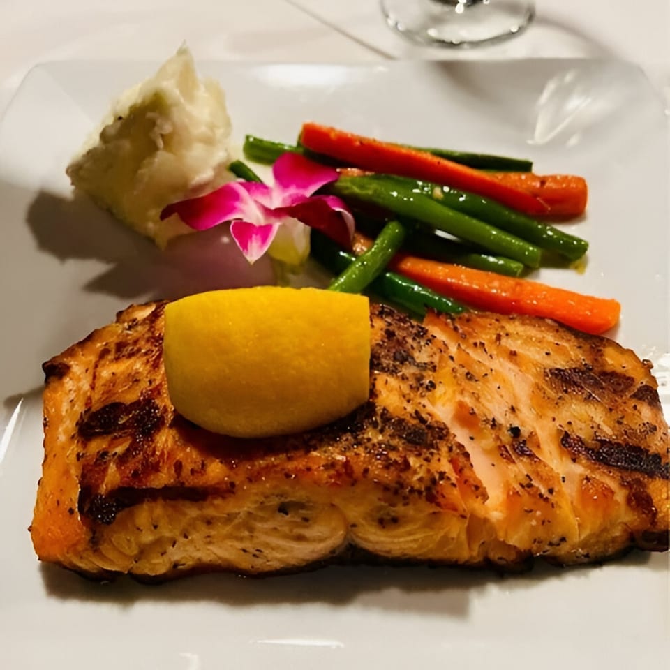 Grilled salmon with a lemon wedge, mashed potatoes, green beans, carrots, and an orchid garnish.
